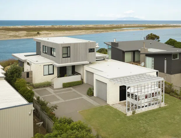 Aerial view of a contemporary coastal residential neighbourhood beside a broad tidal inlet and ocean. The central multi-level home has a boxy form, flat roofs, light weatherboard-style cladding, dark-framed windows, a paved driveway, and a white trellis-covered outdoor structure. Adjacent homes and landscaped yards are visible, with boats on the water in the background.