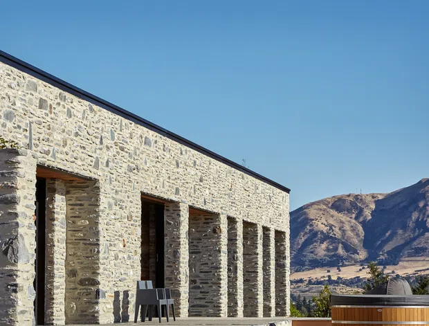 Exterior view of a low, elongated stone building with a dark metal roof edge and a rhythmic colonnade of deep recessed openings. The rough pale masonry facade contrasts with the clear blue sky and rugged mountain landscape beyond. A small portion of an outdoor spa or hot tub is visible in the foreground.