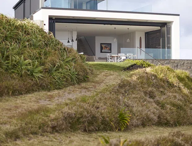 Exterior view of a contemporary two-storey coastal house set into a grassy, landscaped slope. The building has a flat roof with broad overhangs, dark vertical cladding, crisp white rendered walls, extensive glazing, glass balustrades, and a large open ground-level living or dining area with pendant lights and outdoor seating visible inside.