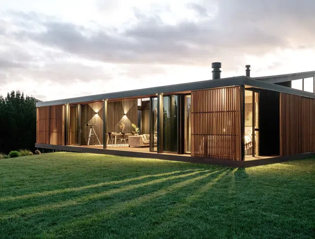 A low-profile modern single-storey home set in a landscaped grassy site. The rectangular building has a flat roof, extensive floor-to-ceiling glazing, dark structural framing, and warm vertical timber screens and cladding. Interior living areas are visible through the glass, with warm evening lighting contrasting against the overcast sky.