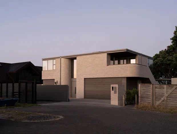 Street-facing view of a contemporary two-storey residence at dusk. The home has a pale rendered masonry facade, dark framed windows, a projecting upper-level form, and an integrated double garage. Timber privacy fencing, a paved driveway, and neighbouring homes frame the suburban setting.
