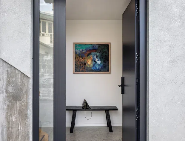 A minimalist contemporary entry viewed from outside through an open black-framed door. The entrance has a broad black metal canopy, narrow glazed sidelight, pale textured exterior wall panels, and a simple interior with a polished concrete or terrazzo-like floor, slim dark console table, small sculptural object, and colourful framed artwork.