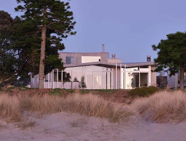 Exterior view of a low-rise contemporary coastal residence set behind natural sand dunes and grasses. The home has a pale, finely detailed facade, extensive glazing, slender vertical posts, a mostly flat roofline, and integrated outdoor-facing living areas. Mature wind-shaped trees frame the property, creating a calm beachfront architectural setting.