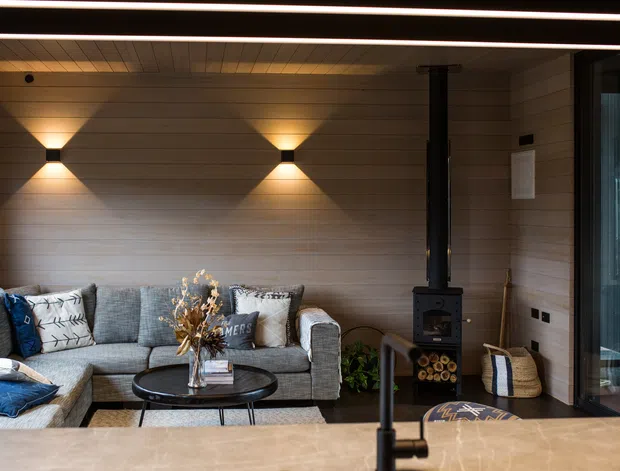 A warm, contemporary living room with a grey sectional sofa arranged around a round black coffee table. The room features horizontal timber wall and ceiling lining, integrated warm wall sconces, a black wood-burning stove with flue, and large glazed doors on the right. A timber-toned benchtop or island edge with a black tap is visible in the foreground, suggesting the living area adjoins an open-plan kitchen. The atmosphere is cozy and refined, with strong linear lighting and natural materials.