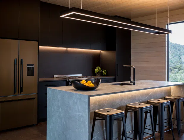 A sophisticated contemporary kitchen with a large stone-clad island and integrated seating for four. Dark, matte cabinetry and a matching refrigerator create a dramatic backdrop, while warm under-cabinet lighting and linear pendant lighting illuminate the island. The island features a dark sink and sculptural black tap, with timber wall panelling and expansive glazing on the right overlooking a green landscape.