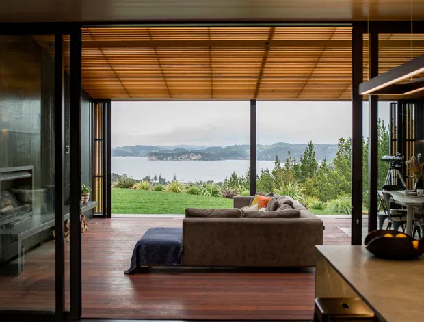 A contemporary open-plan living space with dark timber flooring and exposed timber ceiling slats, opening through large black-framed glazed doors to a landscaped lawn and expansive waterfront view. A brown sofa faces the opening, with a fireplace on the left and a dining area with linear pendant lighting on the right. The room has a warm, refined indoor-outdoor connection and strong horizontal framing.