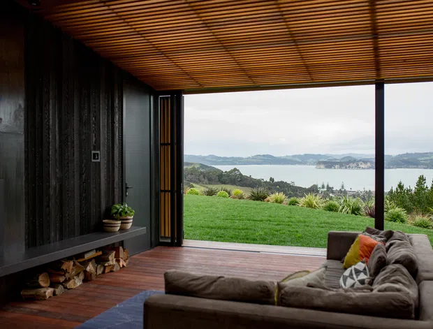 A dark, timber-lined living room opens through wide floor-to-ceiling sliding doors onto a timber deck and lawn, with expansive views across a bay and surrounding hills. The room features a low built-in fireplace wall and firewood storage, a large sectional sofa, warm timber slatted ceiling, and strong indoor–outdoor connection.