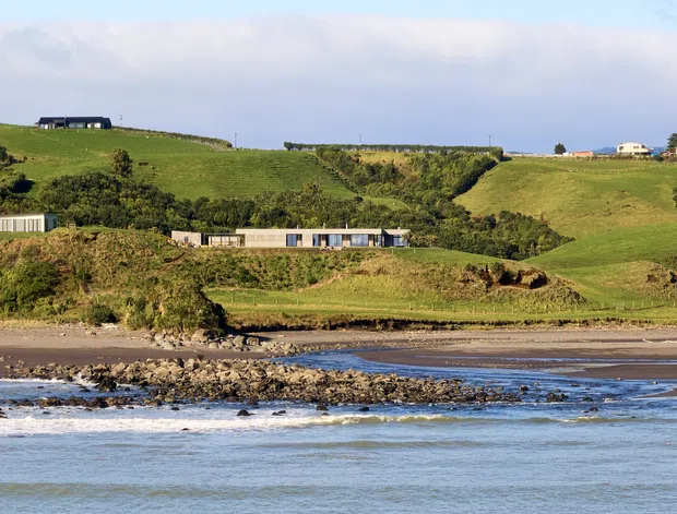 A wide coastal landscape view showing a low, modern residential building integrated into rolling green farmland above a rocky shoreline. Additional rural homes are scattered across the hills, with calm shallow water, beach, and expansive sky in the background.