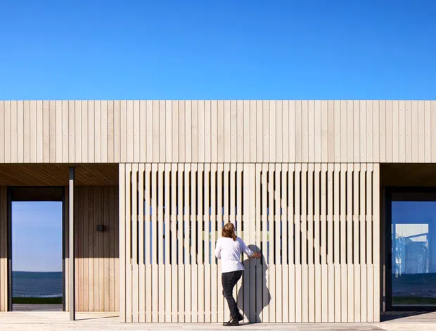 Contemporary single-storey building facade under a clear blue sky, featuring pale vertical timber cladding and a prominent slatted timber privacy screen. Dark-framed glazed openings flank the central screen, with glimpses of an expansive blue horizon visible through the glazing. A person stands at the screen, providing scale.
