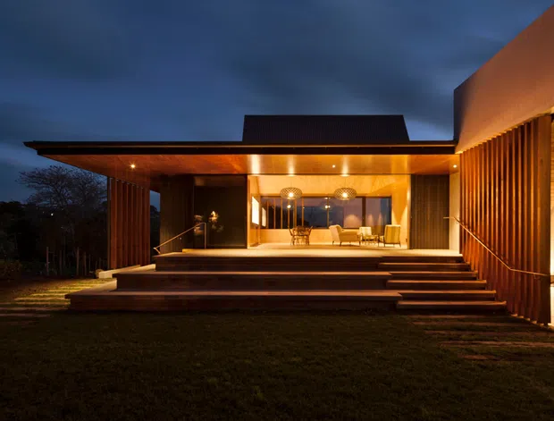 Twilight exterior view of a contemporary, low-slung residence with broad flat roof planes, warm timber-lined soffits, vertical timber screens, and extensive illuminated glazing. A raised timber deck and wide stepped entry terrace lead into a warmly lit open-plan interior, while the dark landscape and sky create a dramatic evening atmosphere.