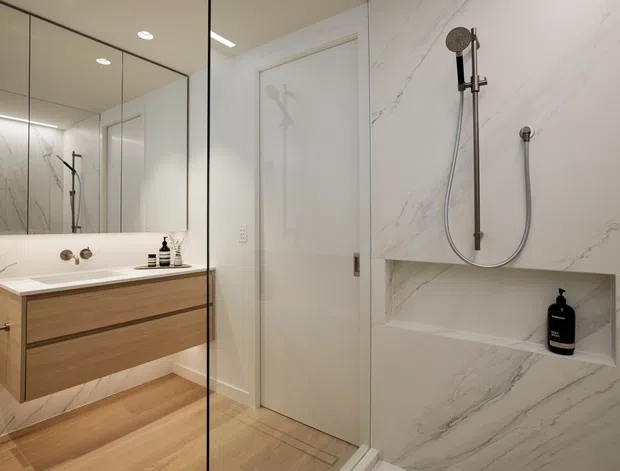 Contemporary minimalist bathroom with a frameless glass shower enclosure, large white marble-look wall tiles with grey veining, a recessed shower niche, wall-mounted brushed metal shower fittings, a floating light-wood vanity, integrated basin, and large mirrored cabinetry. The palette is light, warm, and refined.
