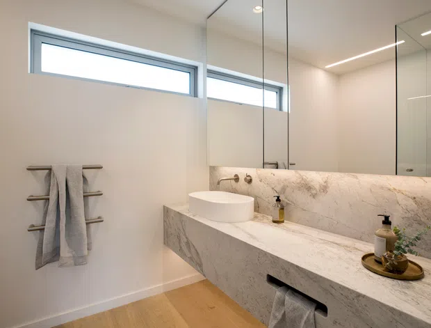 Contemporary bathroom with a floating stone vanity, integrated basin, large mirrored wall cabinets, frameless glass shower enclosure, horizontal clerestory windows, and a heated towel rail. The space uses warm timber flooring, pale walls, and veined light stone for a clean, refined appearance.