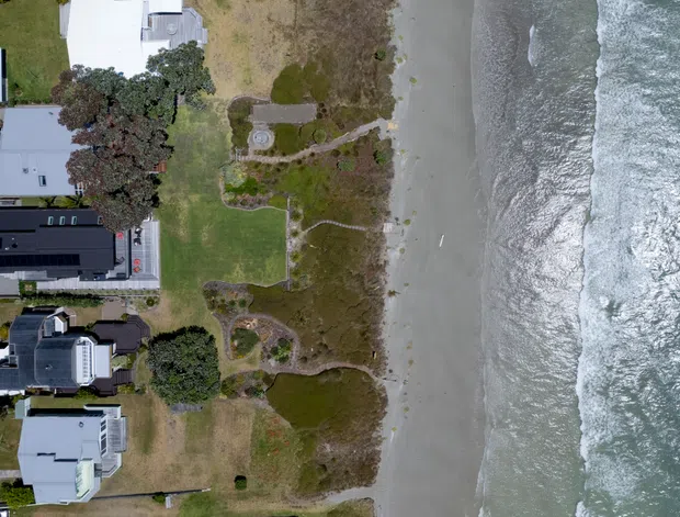 Aerial view of a beachfront residential neighbourhood, showing several detached homes, landscaped gardens, a sandy beach, and ocean waves along the shoreline. One prominent dark-roofed property includes extensive lawn and garden areas directly adjoining the beach.