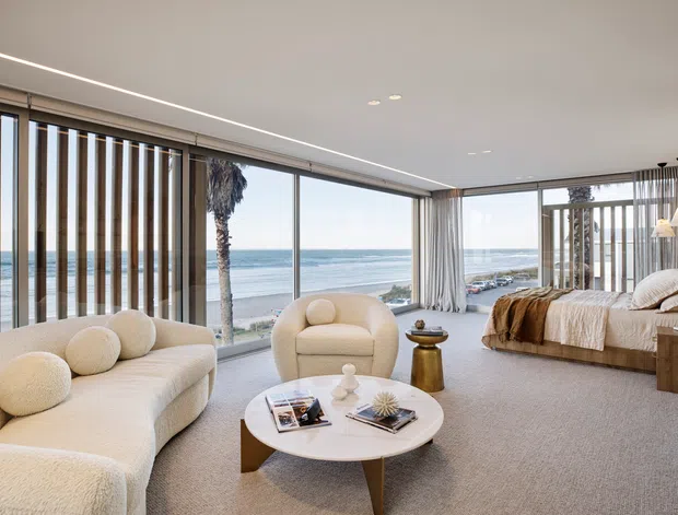 A bright, open-plan coastal living room and bedroom suite with floor-to-ceiling glazing overlooking the ocean. The room features a curved cream sectional sofa, rounded armchair, circular coffee table, neutral carpet, linen bedding, timber bedside cabinetry, sheer curtains, and vertical timber screening elements. The interior has a soft, refined contemporary feel with abundant natural light and a direct beach outlook.