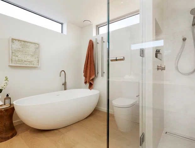 Bright contemporary bathroom with a freestanding white oval bathtub, wall-mounted bath filler, frameless glass shower enclosure, rainfall and handheld shower fittings, and a concealed toilet. Pale timber flooring, white walls, a large horizontal window, and warm wood accents create an airy, refined space.