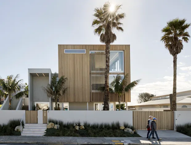 Street-level exterior view of a contemporary two-storey residence with a box-like form, vertical timber cladding, white rendered walls, a projecting upper floor, and large glazed openings screened by vertical timber battens. Palm trees, low coastal-style landscaping, a pedestrian sidewalk, and a driveway are visible in front of the property.