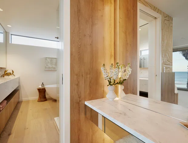 Warm, refined bathroom interior viewed from an adjacent vanity or dressing area. The space features a long timber vanity with a pale stone benchtop, full-height timber wall panelling, a freestanding white bathtub, light timber flooring, mirrored surfaces, and a coastal ocean view through the glazing. A sculptural carved feature panel and neutral finishes add decorative detail.