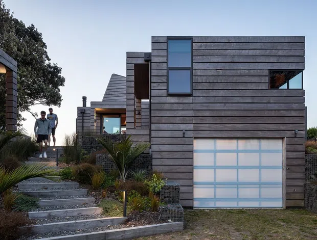 Exterior view of a contemporary multi-level home on a landscaped sloping site. The building has dark horizontal timber-like cladding, strong rectilinear volumes, large glazed openings, and an integrated garage with a translucent paneled door. Stepped concrete paths, native-style planting, gabion retaining walls, and a small group of people are visible.