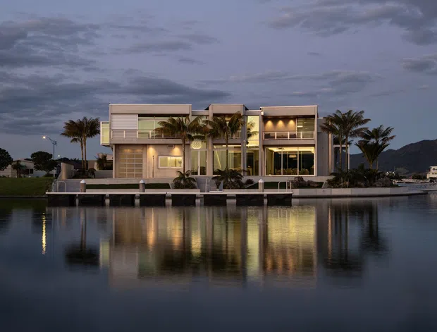 Twilight exterior view of a contemporary multi-storey waterfront residence, with broad glazed openings, pale horizontal cladding, balconies, illuminated interiors, palm trees, and strong reflections across the calm canal or lagoon.
