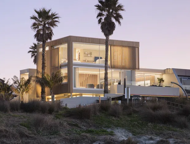 A contemporary multi-level beachfront residence photographed at dusk, with expansive glazing, warm interior lighting, vertical timber screening, clean rectilinear volumes, and a flat roofline. Palm trees and sandy coastal vegetation frame the house, with the beach in the foreground.
