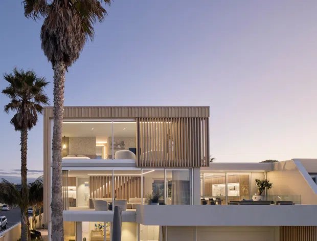 Contemporary multi-level residence photographed at dusk, featuring extensive floor-to-ceiling glazing, white rendered volumes, warm timber vertical screening, broad balconies, and an integrated garage. Tall palm trees frame the street-facing facade, while warmly lit interiors are visible through the glass.