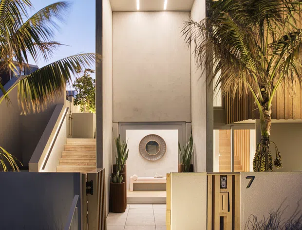 A dusk exterior view of a contemporary residential entry. Stepped tiled stairs lead through an open gate to a covered entrance courtyard, with a tall recessed portico illuminated by linear ceiling lights. The entry includes a built-in bench, circular wall feature, potted plants, palms, a timber-clad upper facade, rendered walls, and a visible house number 7.