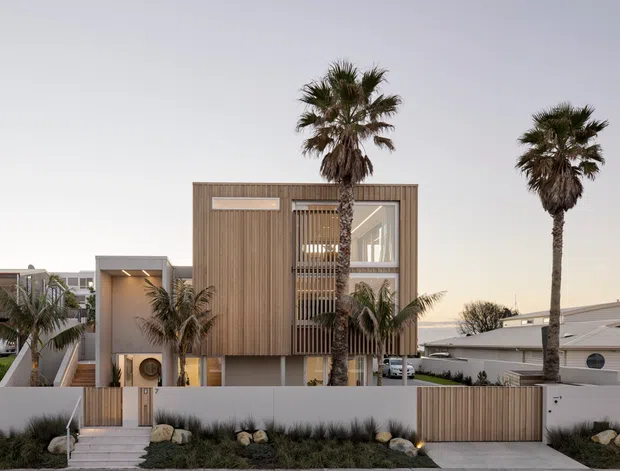 Contemporary multi-level residential facade with vertical timber cladding, concrete elements, screened glazing, and a recessed entry. Tall palm trees, landscaped planting, a timber privacy gate, and neighbouring homes frame the coastal suburban streetscape.