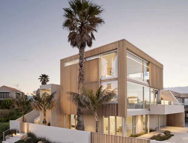 Contemporary multi-storey residential house with a strong timber-clad cubic form, extensive floor-to-ceiling glazing, recessed balconies, and vertical timber screening. The landscaped frontage includes palm trees, low planting, stone accents, and a rendered boundary wall with timber gates. A neighbouring suburban coastal-style setting is visible in the background.