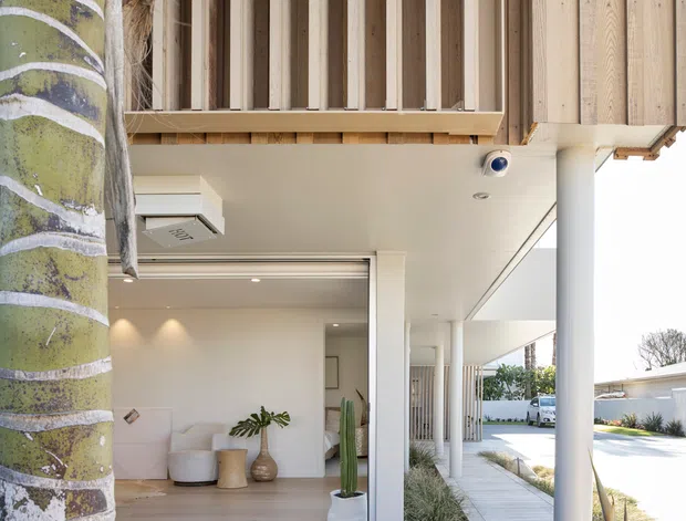 A bright contemporary coastal-style house exterior viewed from a landscaped garden. The image highlights a covered entry or outdoor living area with white cylindrical columns, pale timber decking and cladding, vertical timber screening, large glazed openings, and a recessed interior lounge visible through sliding glass doors. Tropical planting and a palm trunk frame the foreground.