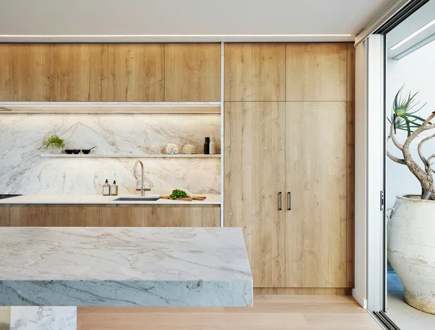 A refined contemporary kitchen with full-height light timber cabinetry, open shelving, and a dramatic veined stone splashback and island benchtop. A stainless-steel sink and tap sit beneath the shelving, while large sliding glass doors open to a balcony or terrace with a potted plant. The palette is warm and natural with clean-lined built-in joinery.