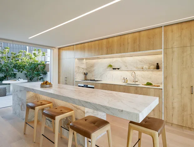 A bright, spacious contemporary kitchen featuring a large waterfall-edge stone island with four leather-upholstered stools, full-height light timber cabinetry, and a marble-look stone splashback with open display shelving. Integrated appliances and minimal linear lighting create a clean, refined interior, while glazing and indoor greenery bring in natural light.