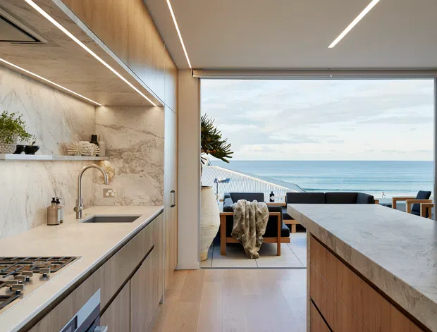 A refined open-plan kitchen overlooking the ocean, with a large stone island and waterfall benchtop, integrated timber cabinetry, marble-look wall surfaces, gas cooktop, and a dining/living area opening onto a seaside terrace. Linear recessed lighting and warm timber finishes create a clean contemporary coastal interior.