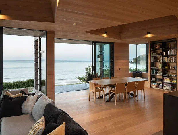 A refined open-plan coastal living and dining space with expansive floor-to-ceiling glazing overlooking the ocean. Warm timber-lined ceilings, walls and cabinetry are paired with timber flooring, a large sectional sofa, a wood dining table with matching chairs, and a dark built-in shelving unit. The room opens toward a bright waterfront outlook and includes integrated recessed and concealed lighting.