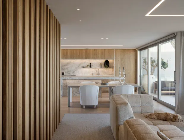 A refined open-plan living and dining area with a warm timber feature screen at left, a light wood and marble kitchen or bar wall at the rear, a round dining table, upholstered dining chairs, and a large neutral modular sofa. Full-height sliding glass doors open to an outdoor area, while linear recessed ceiling lighting and soft curtains create a calm, contemporary interior.