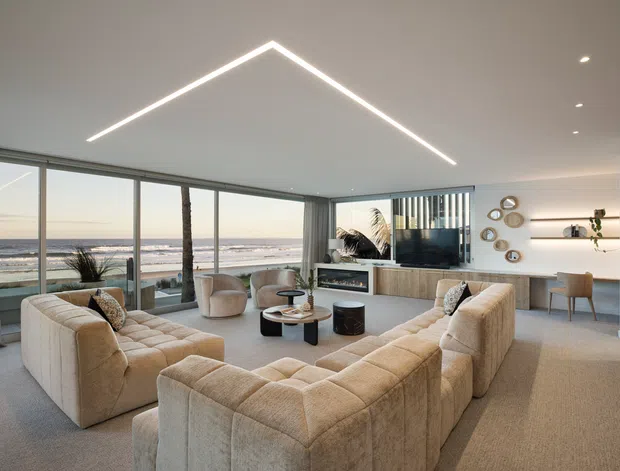 A spacious contemporary living room with a large cream modular sectional arranged around a low coffee table and a built-in fireplace/media unit. Full-height glazing overlooks a beach and ocean, while recessed spotlights, a geometric linear ceiling light, pale timber joinery, and neutral textured finishes create a refined coastal atmosphere.