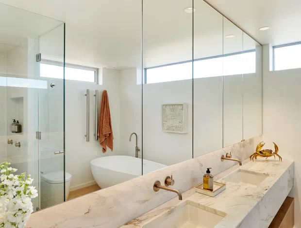 Bright contemporary bathroom featuring a long double vanity with a thick white veined stone benchtop, integrated rectangular basins, timber cabinetry, a freestanding bathtub, glass-enclosed shower, wall-to-wall mirrors, and high horizontal windows. Recessed ceiling lights and warm timber accents create a clean, refined atmosphere.
