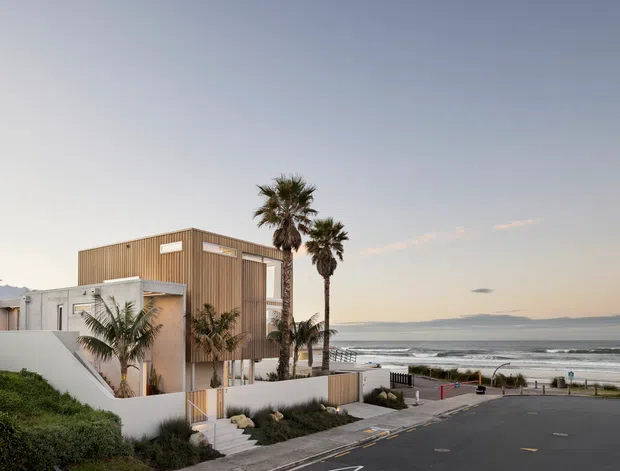 Contemporary multi-level coastal residence with a boxy timber-clad upper volume, white rendered walls, and large glazed openings. The house sits beside a beachfront road, with palm trees, landscaped planting, and ocean waves visible beyond.