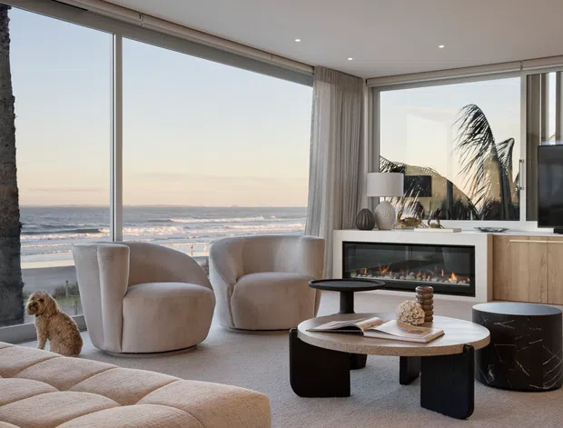 A refined contemporary coastal living room overlooking the ocean, with expansive corner glazing, sheer curtains, curved upholstered lounge chairs, a sculptural coffee table, built-in fireplace, timber cabinetry, and a television. A palm tree and beach are visible outside, while the room is styled in soft neutral tones with rounded forms and a calm, luxurious atmosphere.