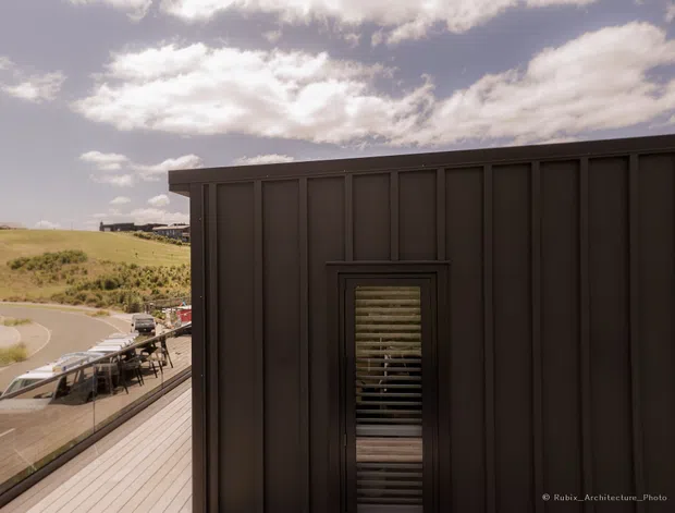 Exterior architectural photograph of a dark, vertically panelled building facade with a narrow louvred window. A timber deck with outdoor dining furniture extends alongside the building, overlooking a gently rolling semi-rural landscape and curving road. The image has a contemporary, rectilinear appearance under a partly cloudy sky.