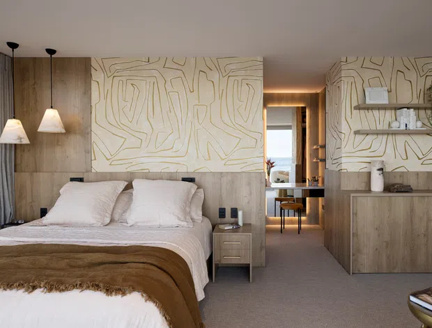 Warm, refined contemporary bedroom with a low upholstered bed, layered neutral bedding, timber wall panelling and built-in cabinetry. A large abstract linear wall treatment spans the headboard wall and continues behind open shelving and storage. Floor-to-ceiling curtains, pendant bedside lights, soft carpet and a view through to a compact desk area create a calm, cohesive interior.