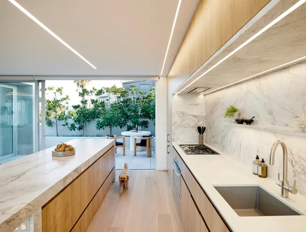 A bright, contemporary galley-style kitchen with a large marble-look island, pale timber cabinetry, integrated linear LED lighting, a stone benchtop and splashback, and a double-bowl undermount sink with brushed metal tapware. Full-height glazing opens toward a small outdoor courtyard or terrace, bringing in abundant natural light.