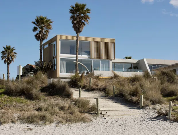 Contemporary coastal house viewed from a sandy beach, featuring a rectilinear form, extensive glazing, warm vertical timber screening, pale cladding, and a timber boardwalk through dune vegetation. Palm trees and beach grasses reinforce the coastal setting.