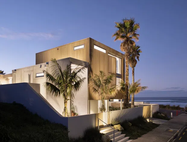 A contemporary multi-storey coastal residence viewed from the street at dusk. The house combines smooth light-coloured rendered walls with a prominent upper volume clad in vertical timber, narrow clerestory-like windows, and angular stepped forms. Palm trees frame the property, while the ocean is visible beyond the road and low boundary walls.