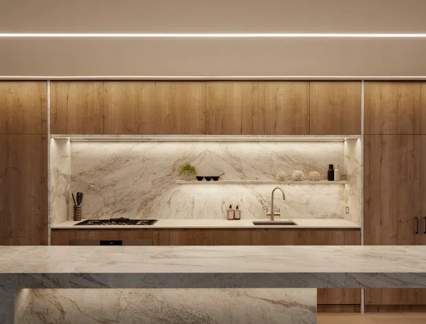 A refined contemporary kitchen featuring full-height warm timber cabinetry, a large pale veined stone island benchtop, and a matching stone splashback with integrated open shelving and concealed lighting. The composition is symmetrical and minimalist, with warm ambient LED lighting and understated fixtures.