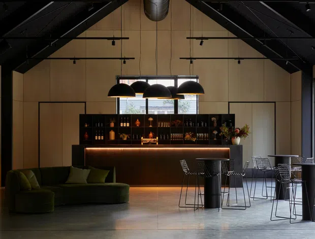 A sophisticated open-plan lounge and home bar with a dark exposed pitched roof structure, warm timber wall panelling, large black-framed windows, and polished concrete flooring. A backlit bar with bottle shelving anchors the room, complemented by sculptural black pendant lights, a deep green curved sofa, and black wire dining tables and chairs. The atmosphere is moody, refined, and contemporary.