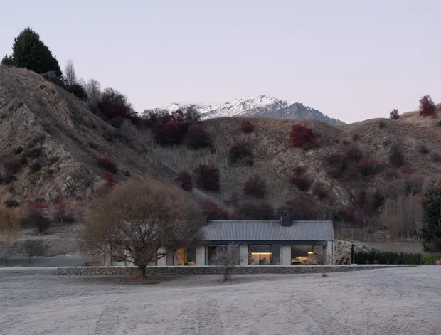 A low, single-storey contemporary residence sits in a rugged rural valley beneath snow-capped mountains. The house has a long, simple form with a dark pitched metal roof, extensive glazing, and stone-clad walls. Frosted ground, sparse deciduous trees, scrub-covered hills, and muted winter colours create a remote alpine setting.