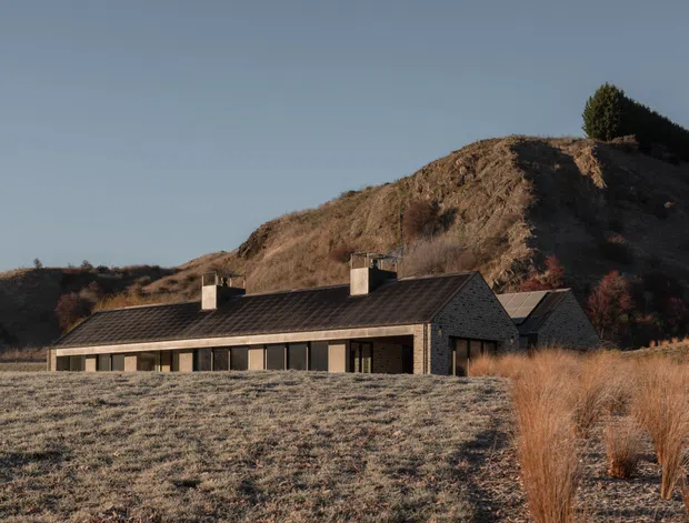 A low-profile contemporary house set into a rural, sloping landscape. The building has dark pitched roofs, exposed grey-brown masonry walls, large horizontal glazing, and multiple chimney volumes. Dry grasses and a steep vegetated hill surround the house, with warm low-angle sunlight creating a quiet, earthy atmosphere.
