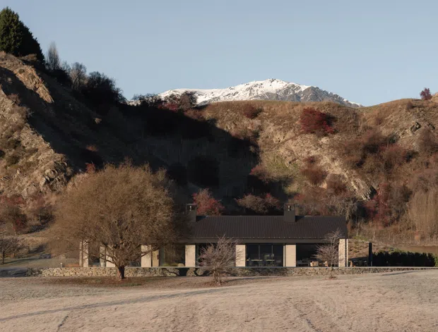 A low, single-storey modern house sits at the base of a rugged, wooded hillside, with a snow-dusted mountain ridge visible behind it. The building has a dark pitched roof, pale walls, extensive dark-framed glazing, and stone-clad sections. A broad unpaved foreground and natural hillside setting dominate the scene.