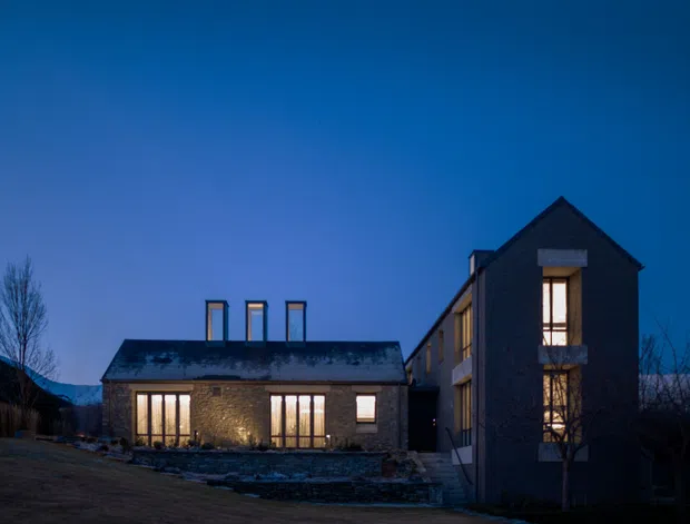 Twilight exterior view of a contemporary multi-building residence with dark masonry cladding, pitched roofs, tall illuminated windows, and a low connecting wing. The property sits against a mountainous landscape with a landscaped foreground.