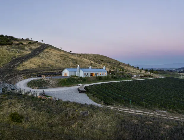 Aerial view of a rural vineyard estate at dusk, showing a long, low white farmhouse or winery building set on a sloping hillside. Gravel access roads, cultivated vine rows, fencing, sparse grassland, and distant coastal or valley scenery surround the property.